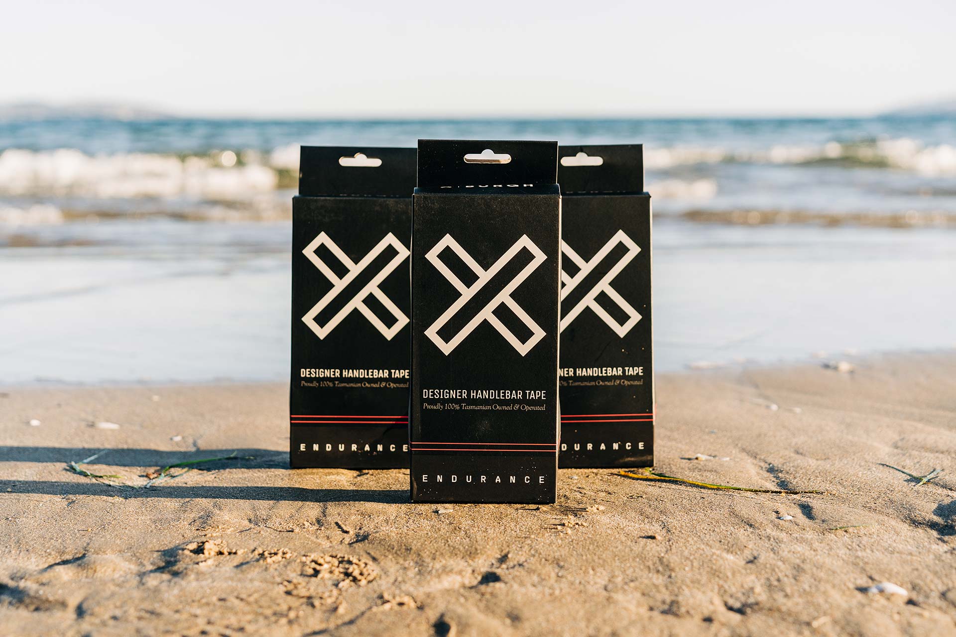 Three boxes of Burgh Bar Tape sitting on the beach with the waves in the background