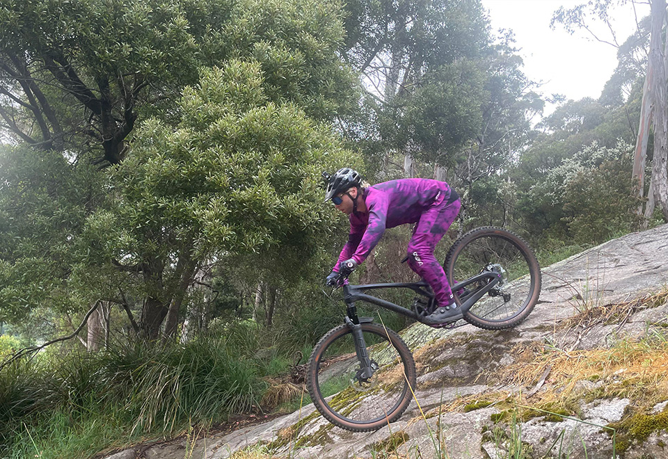 Patrick, in bright purple MTB gear, rides down a rock on his mountain bike.